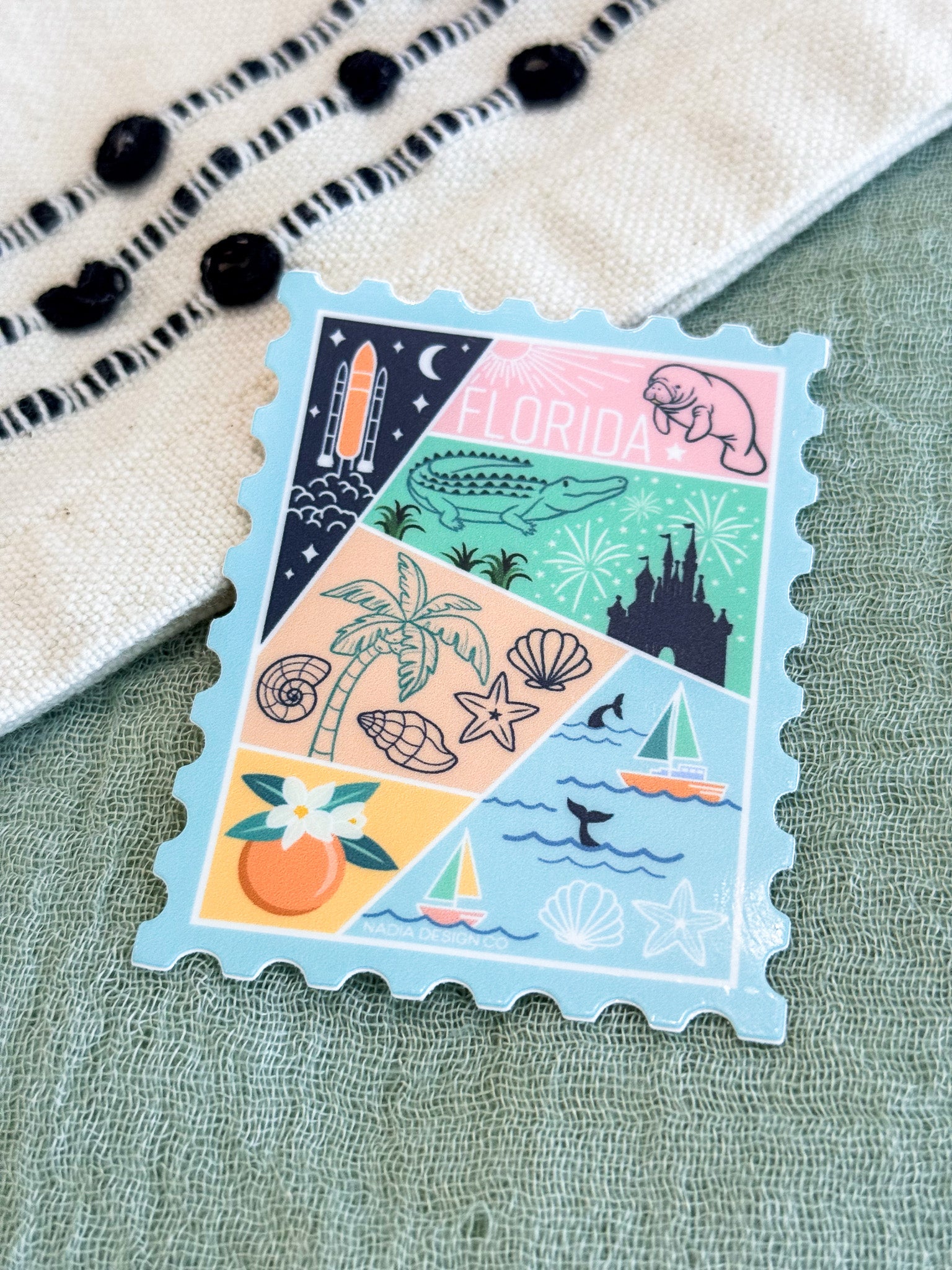 A rectangular postage-stamp-style sticker or decal with scalloped edges, featuring a pastel Florida-themed collage of palm trees, shells, a manatee, an alligator, a rocket, a castle, sailboats, and the word "FLORIDA." It is photographed on a textured light green surface with part of a cream woven textile and black beads visible at the top left.