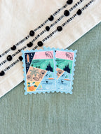 Two Florida postage stamp-style stickers with a multicolor beach and landscape collage design, placed on a sage green textured surface next to a cream embroidered fabric.