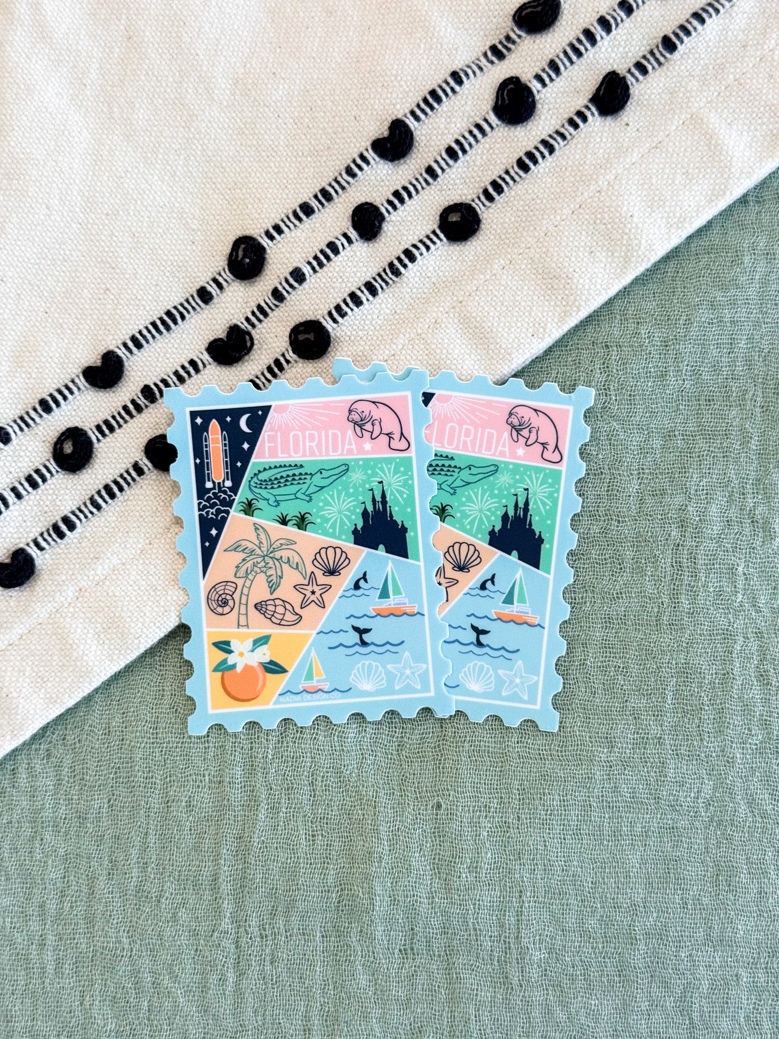 Two Florida postage stamp-style stickers with a multicolor beach and landscape collage design, placed on a sage green textured surface next to a cream embroidered fabric.