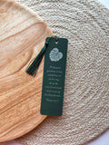 A green bookmark with a tassel featuring a monstera leaf design and a Bible verse, placed on a wooden surface and a beige mat.