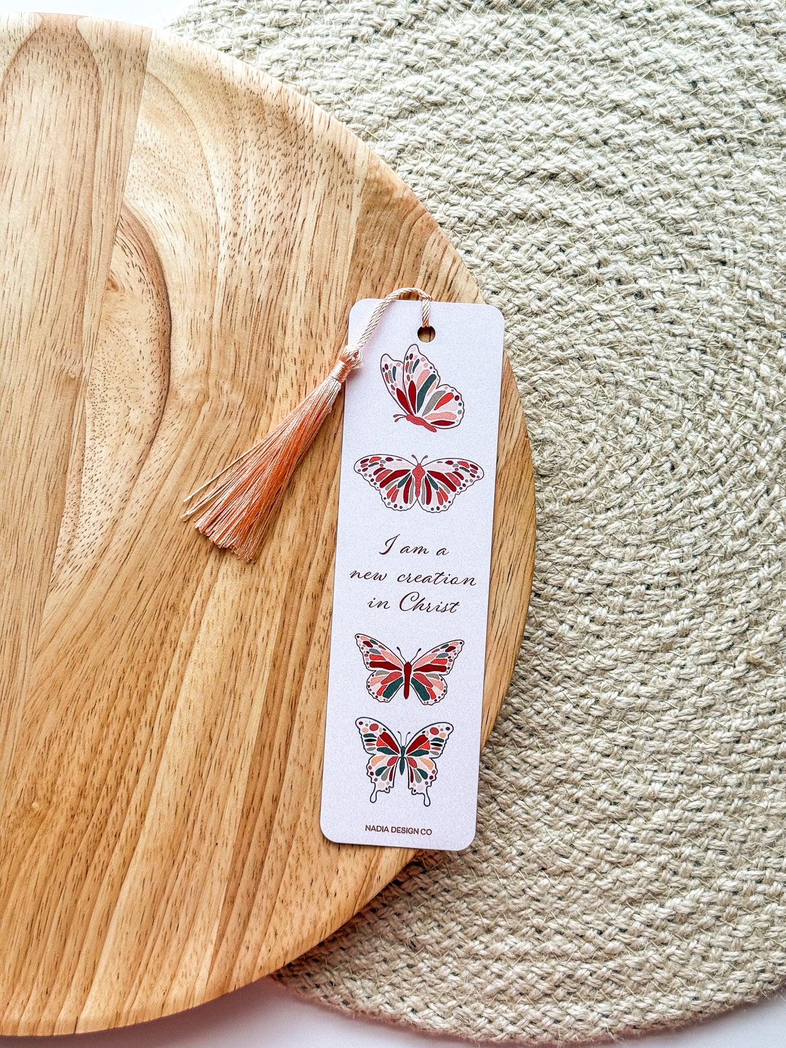 A bookmark with a white background featuring red and green butterfly illustrations and text that reads 'I am a new creation in Christ'. with peach tassel. The bookmark is placed on a wooden surface with a patterned fabric beside it.