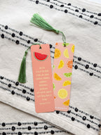 A pink bookmark with a tassel featuring a various fruit design and a Galatians 5:22-23 Bible verse, placed on a open bible. 