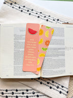 A pink bookmark with a tassel featuring a various fruit design and a Galatians 5:22-23 Bible verse, placed on a open bible. 