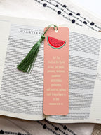 A pink bookmark with a tassel featuring a various fruit design and a Galatians 5:22-23 Bible verse, placed on a open bible. 