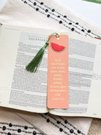 A pink bookmark with a green tassel featuring a various fruit design and a Galatians 5:22-23 Bible verse, placed on a open bible. 