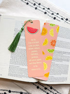 A pink bookmark with a tassel featuring a various fruit design and a Galatians 5:22-23 Bible verse, placed on a open bible. 