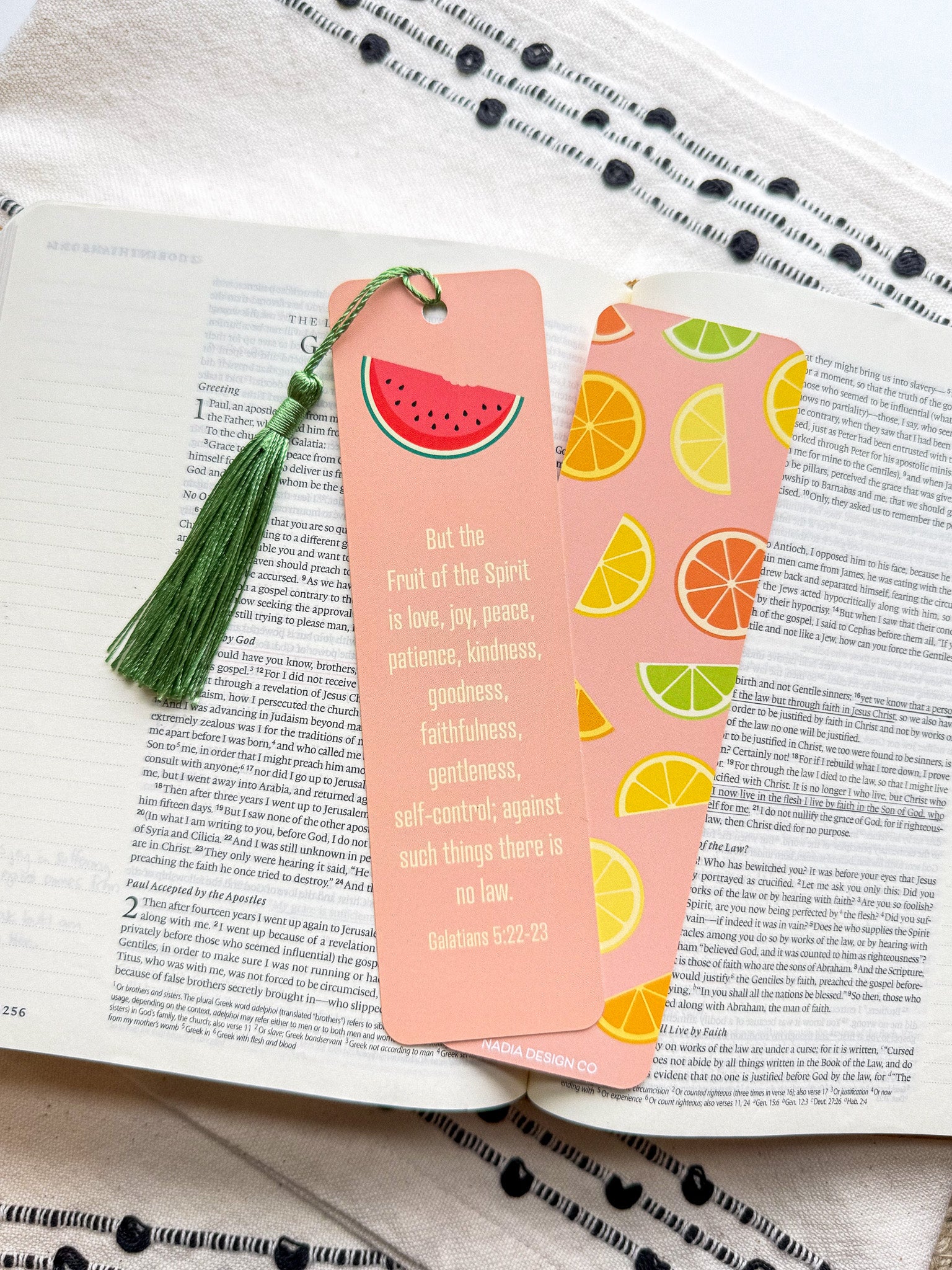 A pink bookmark with a tassel featuring a various fruit design and a Galatians 5:22-23 Bible verse, placed on a open bible. 