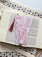 A bookmark with a floral design and a verse from the Bible John 15:12-13, held in a hand. with tassel. The bookmark has a pearl paper finish with rounded corners.