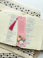 Two pink bookmarks with floral illustrations and a Bible verse written on them with a pink tassel, placed on a bible.