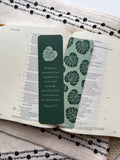 A green bookmark with a tassel featuring a monstera leaf design and a Bible verse, placed on a wooden surface and a beige mat.
