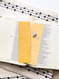 A yellow bookmark with a bee illustration and honeycomb pattern, titled 'Proverbs 16:24 Bible Verse', placed on an open book.