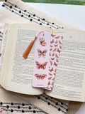 A bookmark with a double-sided illustration of butterflies, printed on pearl paper with a subtle shimmer, placed on an open book.