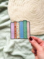 Colorful book-shaped bookmarks held by a hand against a soft fabric background
