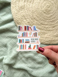 Sticker of a bookshelf with books and 'Read More Books' text held by a hand on a textured fabric background.
