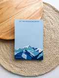 A notepad with a colorful mountain illustration on the cover, titled 'LET THE ADVENTURE BEGIN' placed on a woven pad on top of a wooden surface.