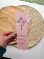 Pink bookmark with a cross and peony floral design held by a hand, with a wooden and woven mat in the background.