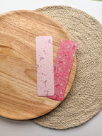Two pink cherry blossom illustrated floral bookmarks on a wooden and woven surface