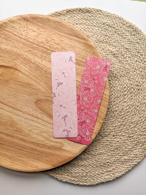 Two pink cherry blossom illustrated floral bookmarks on a wooden and woven surface