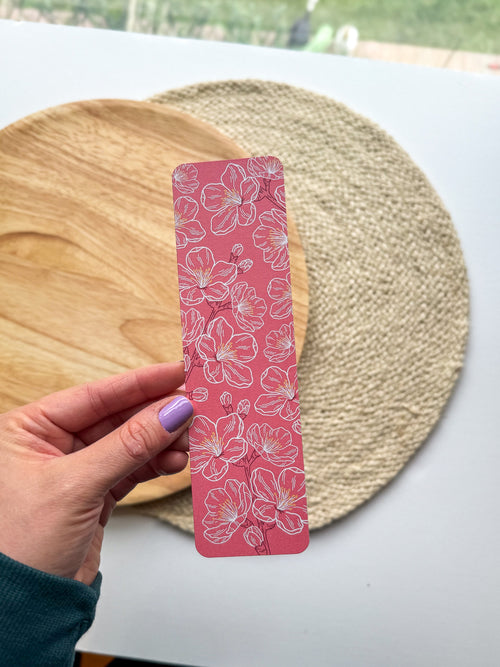 hand holding pink cherry blossom illustrated floral bookmarks on a wooden and woven surface