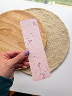 hand holding pink cherry blossom illustrated floral bookmarks on a wooden and woven surface