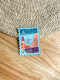 A colorful postage stamp-shaped magnet with an illustration of Rhode Island landmarks, placed on a wooden surface.