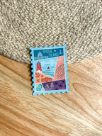 A colorful postage stamp-shaped magnet with an illustration of Rhode Island landmarks, placed on a wooden surface.