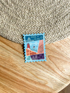 A colorful postage stamp-shaped magnet with an illustration of Rhode Island landmarks, placed on a wooden surface.