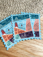 A colorful postage stamp-shaped magnet with an illustration of Rhode Island landmarks, placed on a wooden surface.