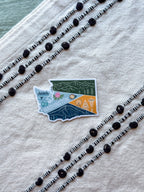 Sticker of a stylized theme of Washington state on a textured fabric background with decorative black and white beads.