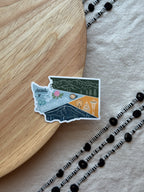 Sticker of Washington state on a wooden surface with decorative elements.