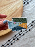 Sticker of Washington state held by a hand on a wooden surface with a patterned fabric background.