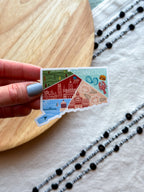 Hand holding a Connecticut-shaped sticker with various icons on a wooden surface.