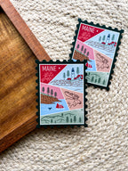 A magnet shaped like a postage stamp with a vibrant paisley-like border, featuring an illustration of various Maine-related items such as a lighthouse, trees, and a lobster boat.