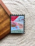 A magnet shaped like a postage stamp with a vibrant paisley-like border, featuring an illustration of various Maine-related items such as a lighthouse, trees, and a lobster boat.