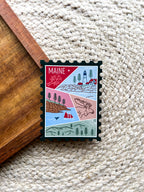A magnet shaped like a postage stamp with a vibrant paisley-like border, featuring an illustration of various Maine-related items such as a lighthouse, trees, and a lobster boat.
