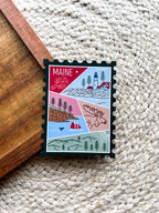 A magnet shaped like a postage stamp with a vibrant paisley-like border, featuring an illustration of various Maine-related items such as a lighthouse, trees, and a lobster boat.