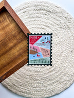 A magnet shaped like a postage stamp with a vibrant paisley-like border, featuring an illustration of various Maine-related items such as a lighthouse, trees, and a lobster boat.