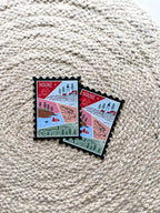 A magnet shaped like a postage stamp with a vibrant paisley-like border, featuring an illustration of various Maine-related items such as a lighthouse, trees, and a lobster boat.