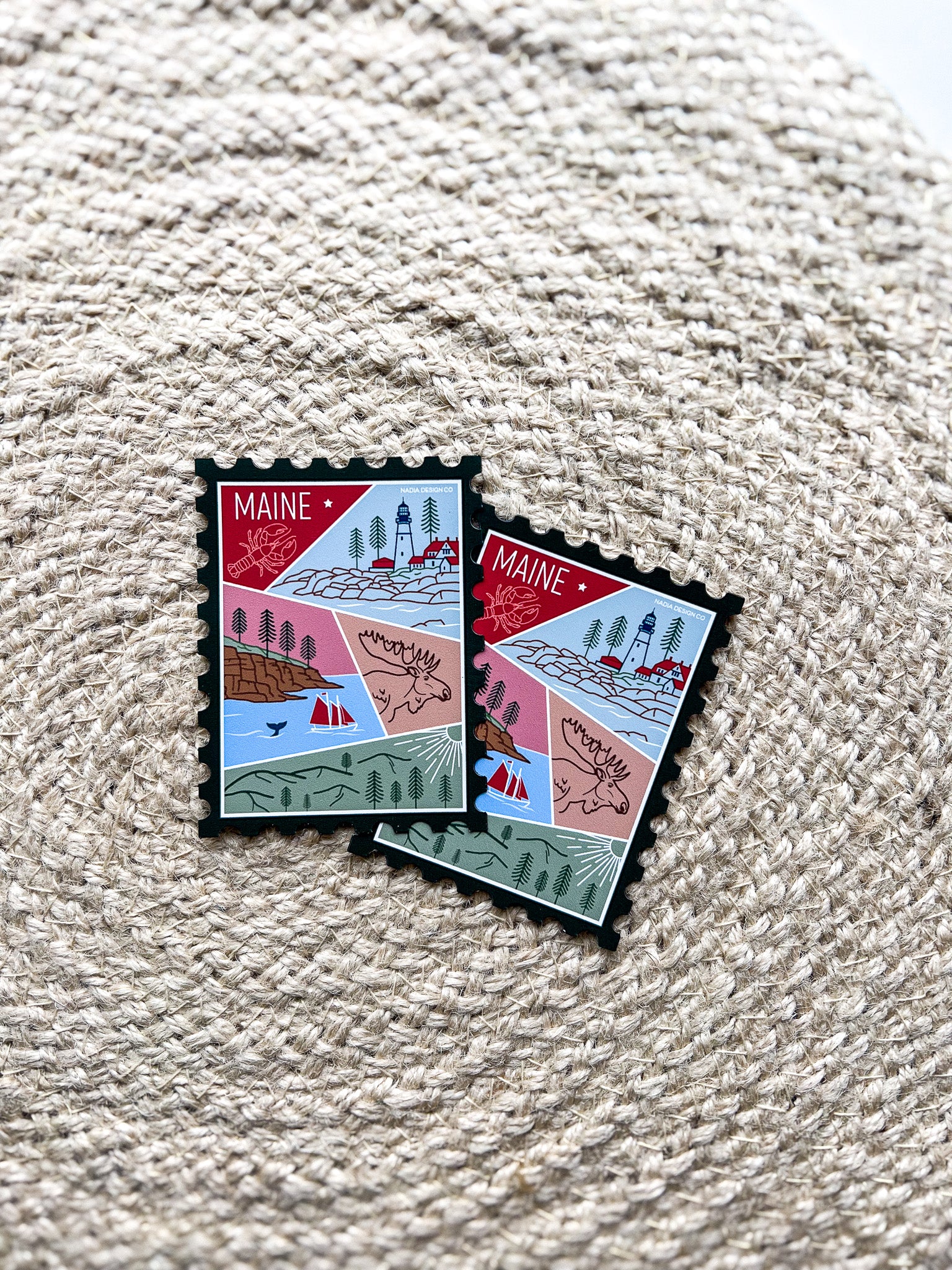 A magnet shaped like a postage stamp with a vibrant paisley-like border, featuring an illustration of various Maine-related items such as a lighthouse, trees, and a lobster boat.