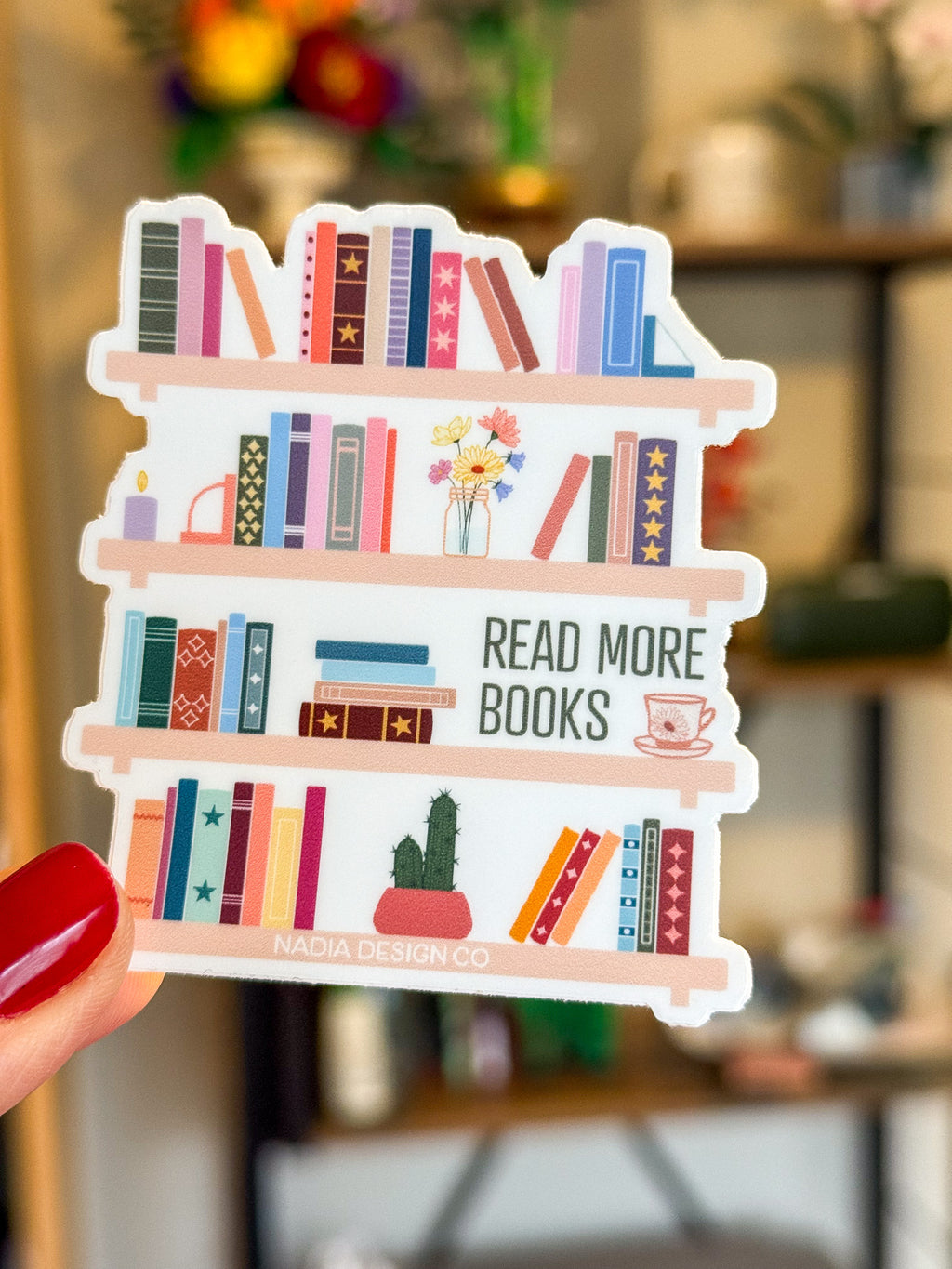 Sticker of a bookshelf with books and 'Read More Books' text, held in front of a blurred bookshelf background.