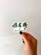 A vinyl sticker with a green and white illustration of various plants in pots, and the text 'Keep Growing!' held by a hand