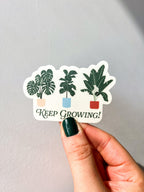 A vinyl sticker with a green and white illustration of various plants in pots, and the text 'Keep Growing!' held up by a hand