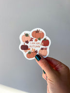 A vinyl sticker with a quote 'I am an Autumn' surrounded by illustrations of pumpkins and autumn leaves held up by a hand