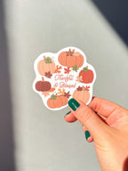 A vinyl sticker with a design of pumpkins, leaves, and the text 'THANKFUL & BLESSED' hand hold it up