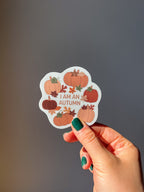 A vinyl sticker with a quote 'I am an Autumn' surrounded by illustrations of pumpkins and autumn leaves held up by a hand