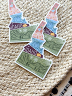 sticker of Idaho with scenic illustrations stamp design held by a hand on a textured fabric background