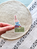 sticker of Idaho with scenic illustrations stamp design held by a hand on a textured fabric background
