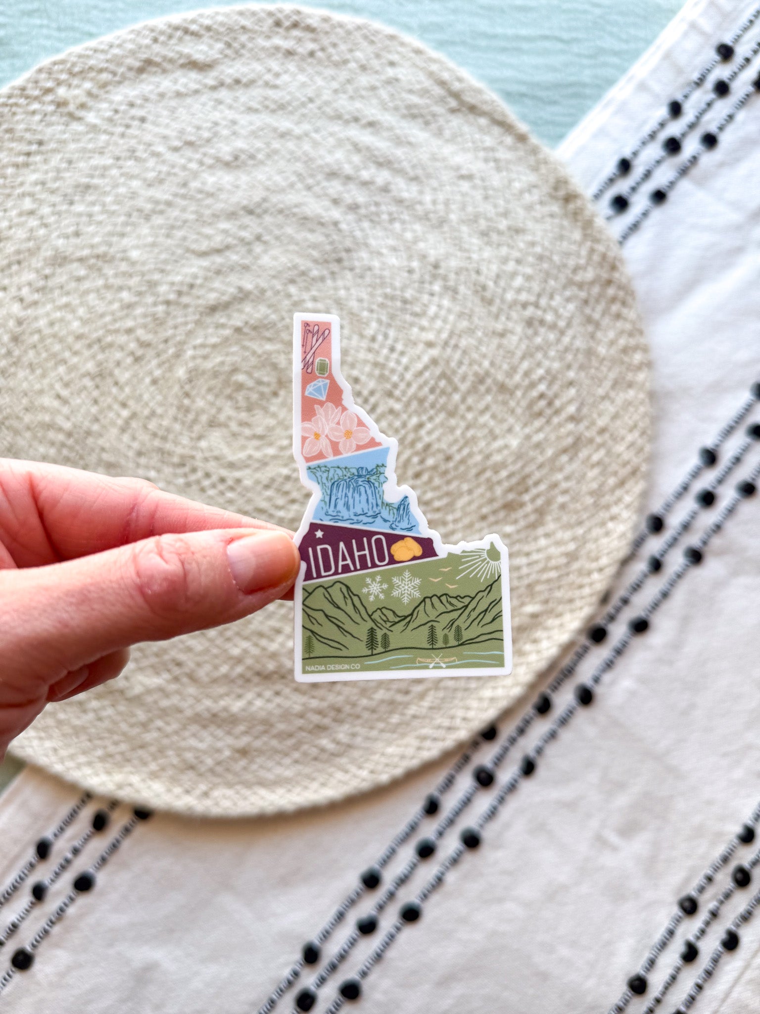 sticker of Idaho with scenic illustrations stamp design held by a hand on a textured fabric background