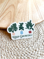 A vinyl sticker with a green and white illustration of various plants in pots, and the text 'Keep Growing!' on a woven mat.