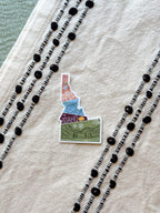 sticker of Idaho with scenic illustrations stamp design held by a hand on a textured fabric background